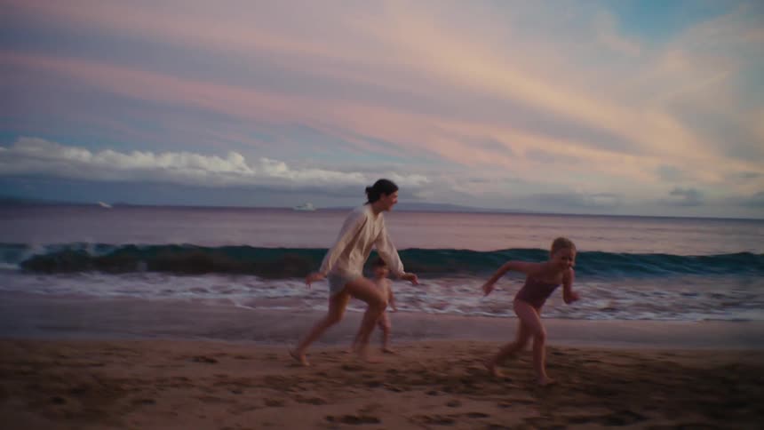 A woman chases a group of children around a sandy beach in the evening