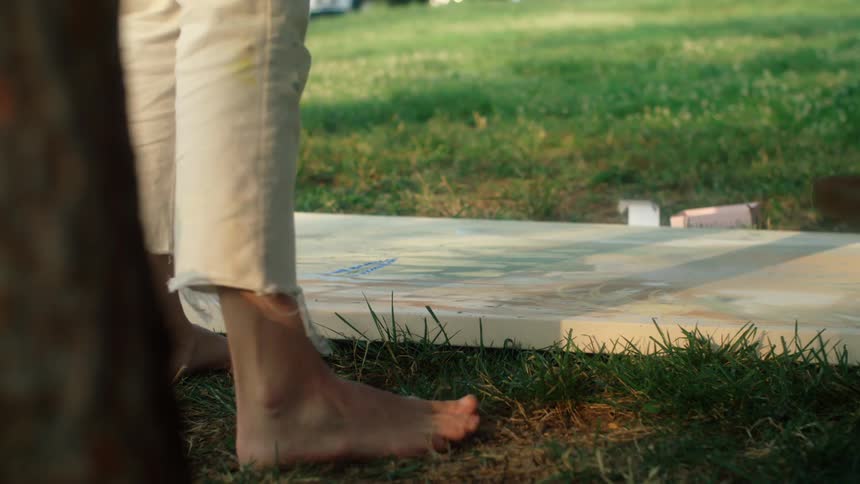 Painter with white pants holding a stick walks barefoot around canvas laying on green lawn