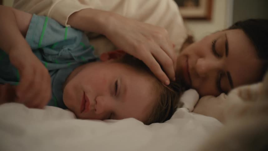 Mother in white sweatshirt strokes toddler sons head and face lying on bed
