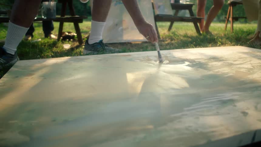 Painter with white socks and sneakers moves brush across the canvas laying on green lawn