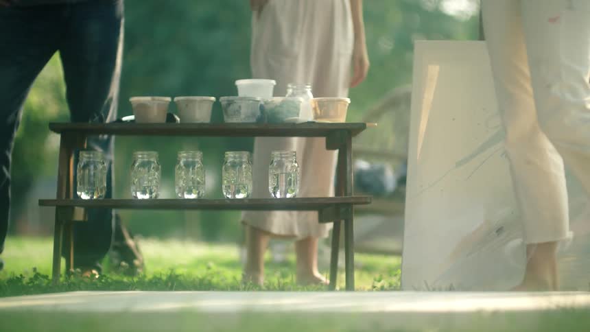 A wood stand in a garden with several empty jars and several boxes of paint and a painting leaning on a tree as three people move between the two