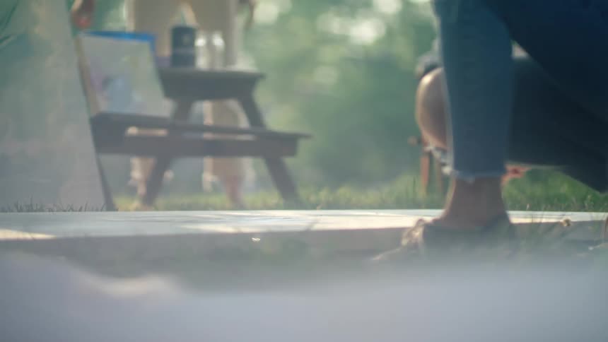 A woman is knelt down in the grass painting on a large canvas