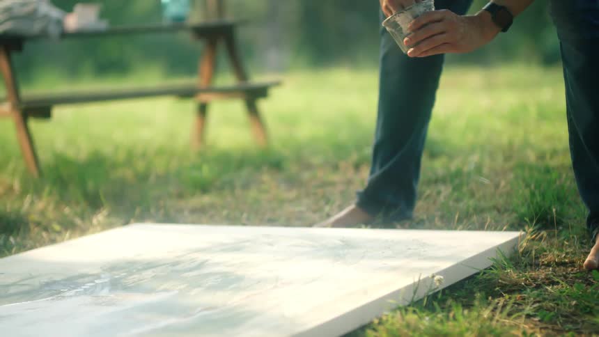 A man speckles paint over a canvas on a grassy ground