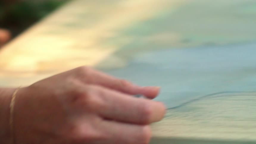 The painter wearing a white shirt hat and pants delicately moves their brush across the canvas as they lay on the lush green grass