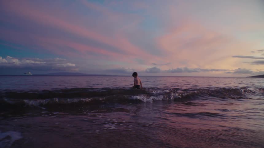 A young boy is on a beach in the water up to his waist when a wave passes him and he looks down