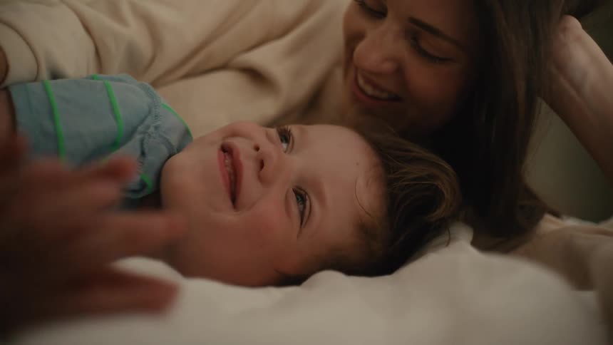 A woman hugs a young boy lying on a bed as they both talk smile and laugh
