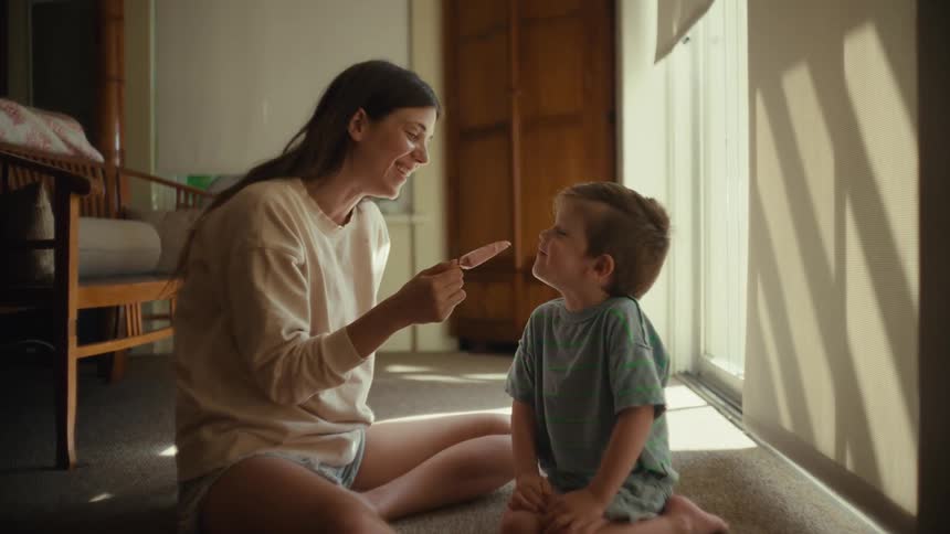 A woman and a boy are sitting cross legged on a living room floor smiling laughing and sharing ice cream