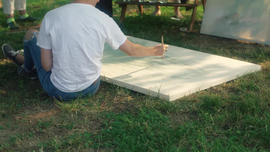 Artist sits on ground painting canvas as spectators watch