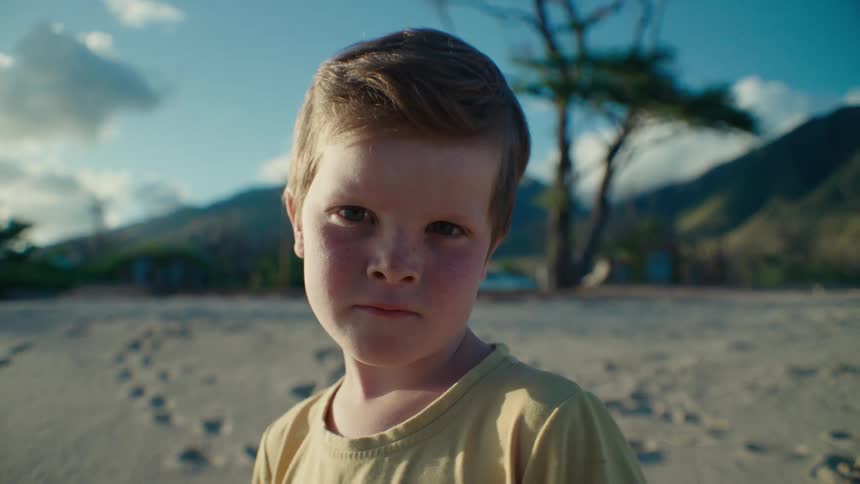 Boy stands on sand strewn with footprints as traffic passes on road between beach and mountains