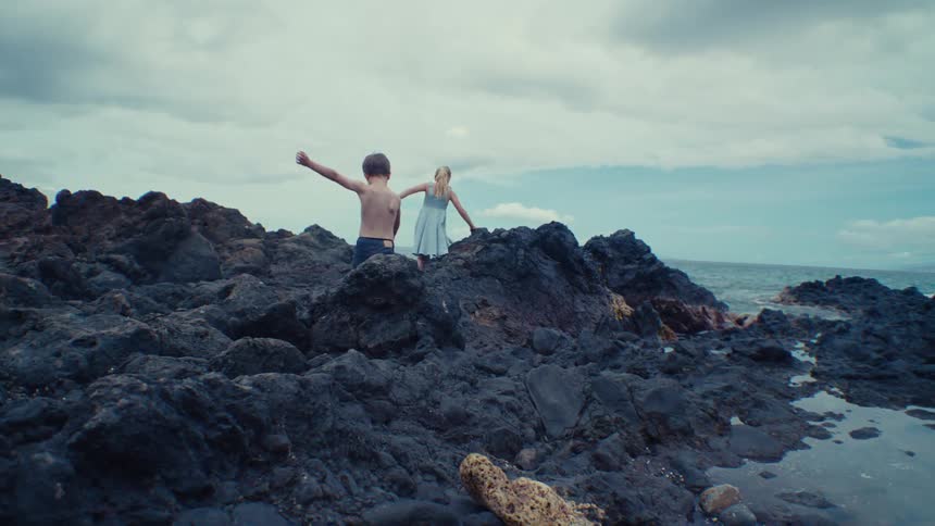 A young girl and a male toddler walk over rocks on a beach