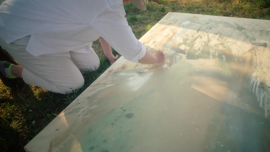 The painter wearing a white shirt hat and pants delicately moves their brush across the canvas as they lay on the lush green grass