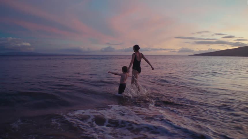 A woman holds her toddlers hand as they both run into the water on a beach