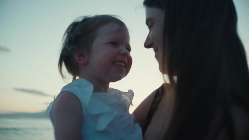 A woman is holding her baby girl in her arms on a beach and they give each other an eskimo kiss