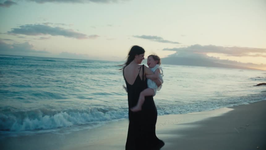 A woman on a beach plays with her baby girl by holding her in her lap and spinning fast as they both laugh