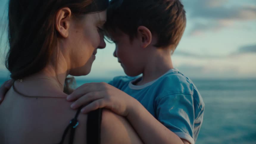 Mother with dark hair and black top holds toddler son on the beach near the ocean at sunset