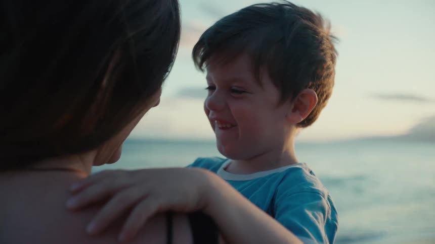 A woman is holding up her young sun as they smile and make faces at each other on a beach