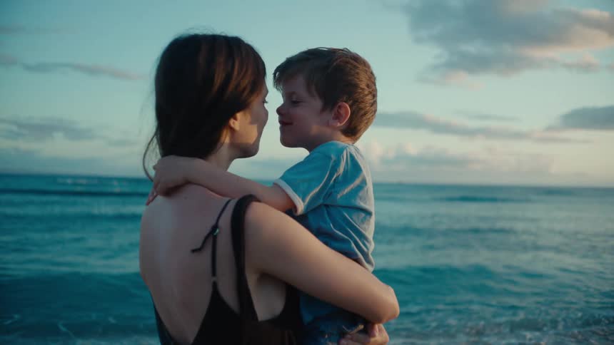 A woman standing on a beach is holding her toddler done in her lap as they smile and eskimo kiss each other