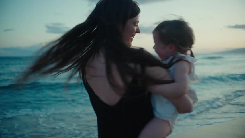 A woman is holding a baby girl in her lap at a beach and starts spinning around to make the baby laugh