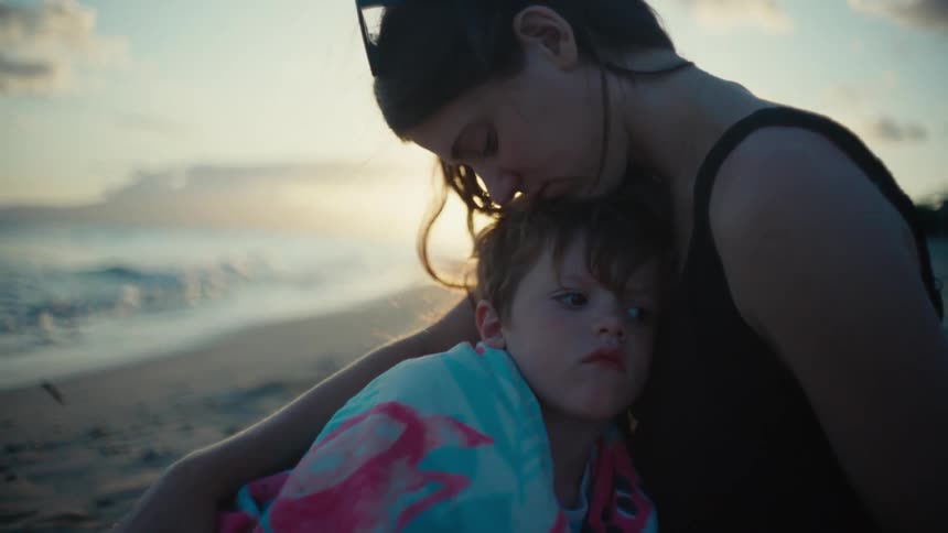 A woman is sitting on a beach holding her toddler son in her arms with a towel wrapped around him and kisses his forehead