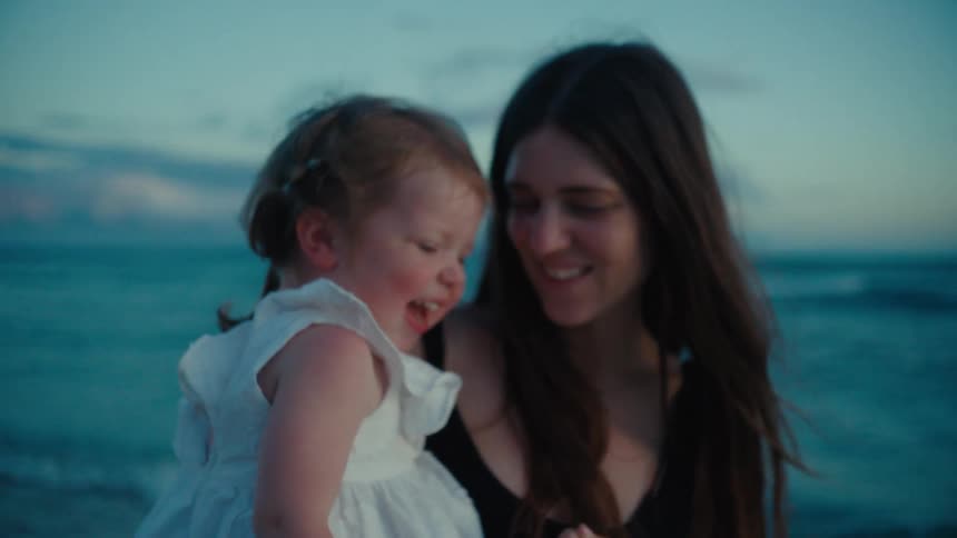 Mother in black dress holds daughter in her arms wearing white dress on the beach at dusk near the ocean