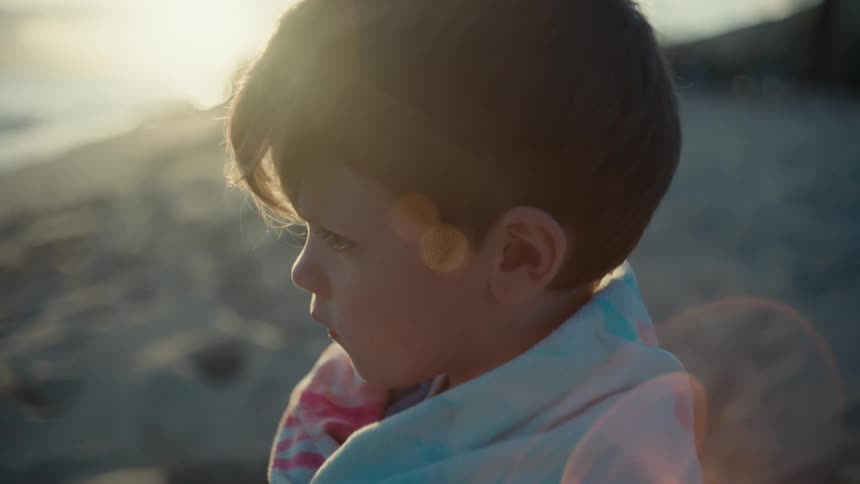 Boy stands wrapped on beach as sun glares across water