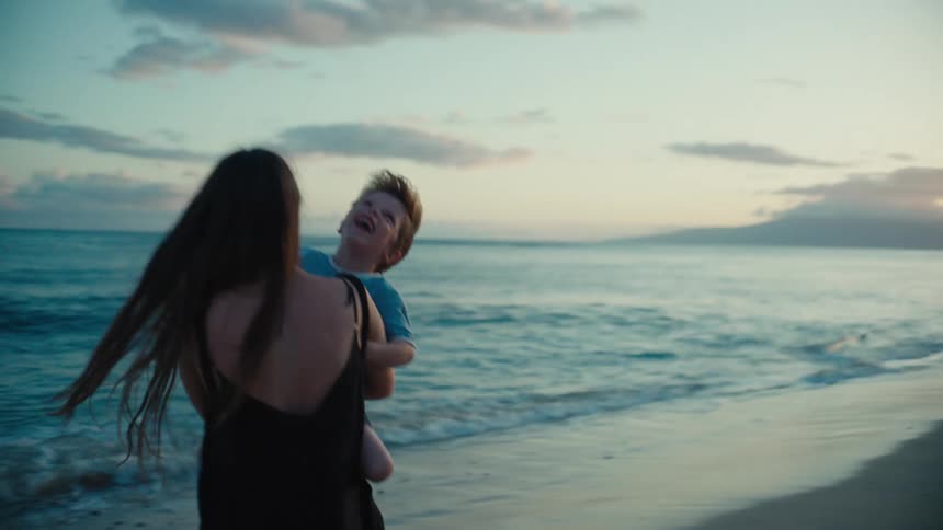 Mother and son both smile as mother holds child and spins on beach