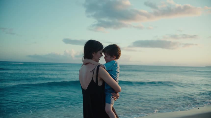 Mother with black dress holds toddler son kissing on the lips on the beach near the ocean