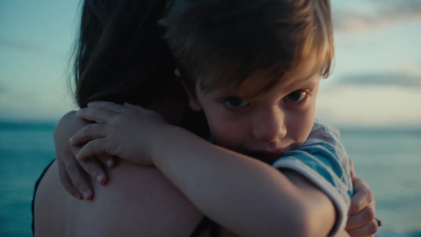 A woman on a beach holds up her toddler son in her arms and they hug