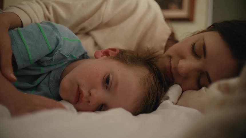 A woman is helping her son fall asleep as she strokes his arm kisses his forehead and sings to him