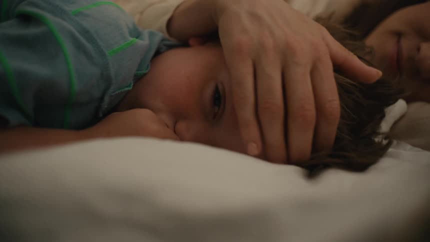 A mother and her toddler son lay tired on a bed as she sweeps hair from his forehead