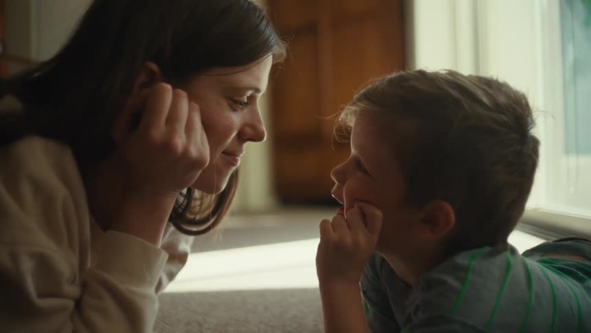 A woman and a toddler are face to face lying on the floor their chins on their hands talking and smiling