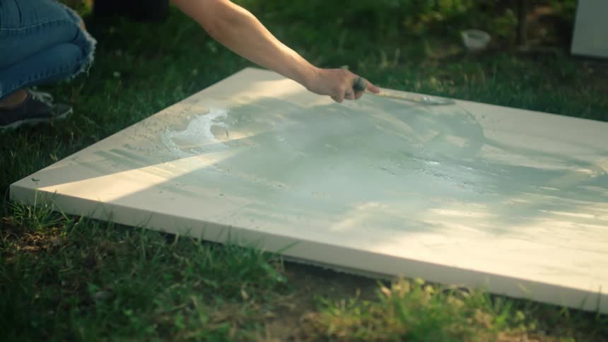 Woman crouches and applies paint to canvas on grass as another person moves and casts shadow onto it