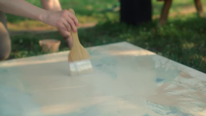 Woman with pants and gray t shirt moves brush across big canvas laying on lawn