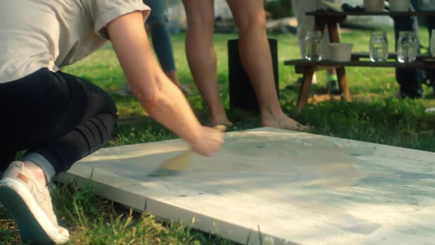 A table top is lying on a grass floor in a picnic area as a man paints it with a brush and a woman looks at him