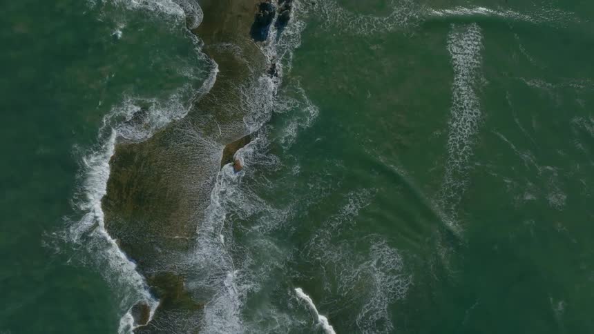 The sea engulfs a small sandy peninsula as waves crash into it