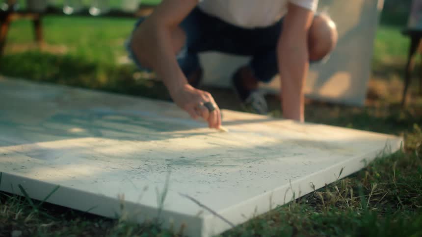 A woman draws with a crayon on a canvas lying on a grassy ground