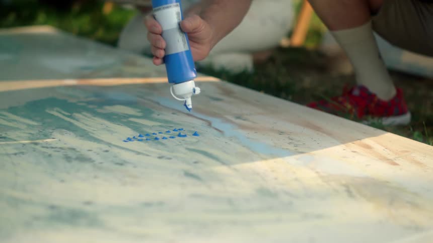 Artist crouches to apply paint from bottle onto canvas lying on the ground