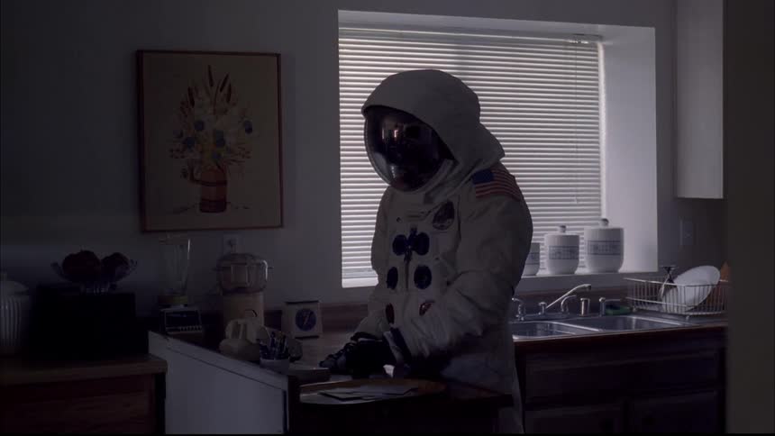 Person wearing astronaut spacesuit stands in kitchen and presses answering machine button