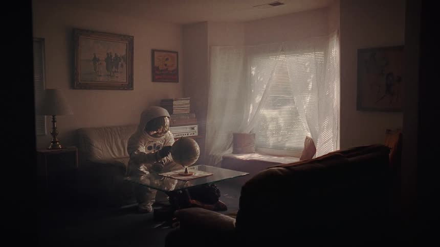 Person wearing astronaut spacesuit sits on loveseat in dim living room and touches globe