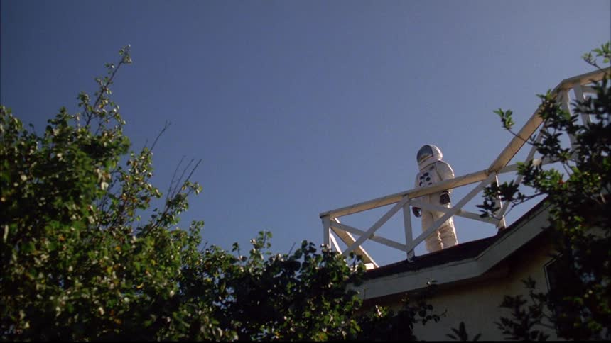 Person wearing astronaut suit stands on house roof near railing