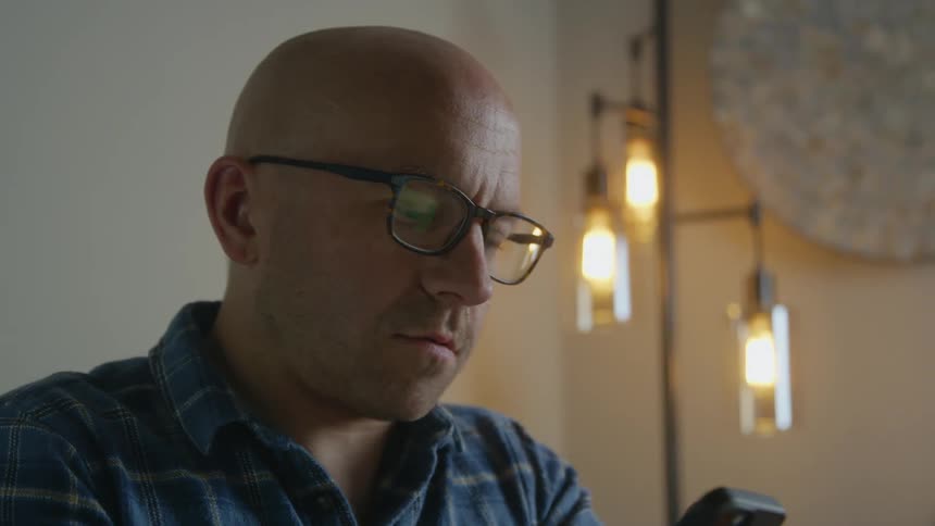 Bald man answers takes call while sitting in room near floor lamp