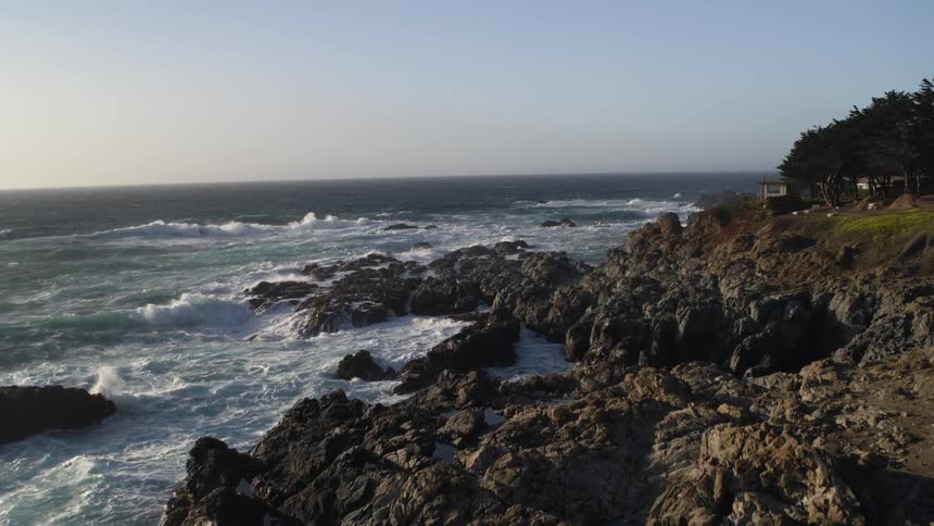 Waves crash against rocky coast near house under a clear sky