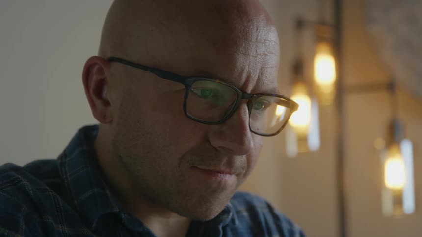 Bald man with glasses and blue shirt uses laptop shaking his head