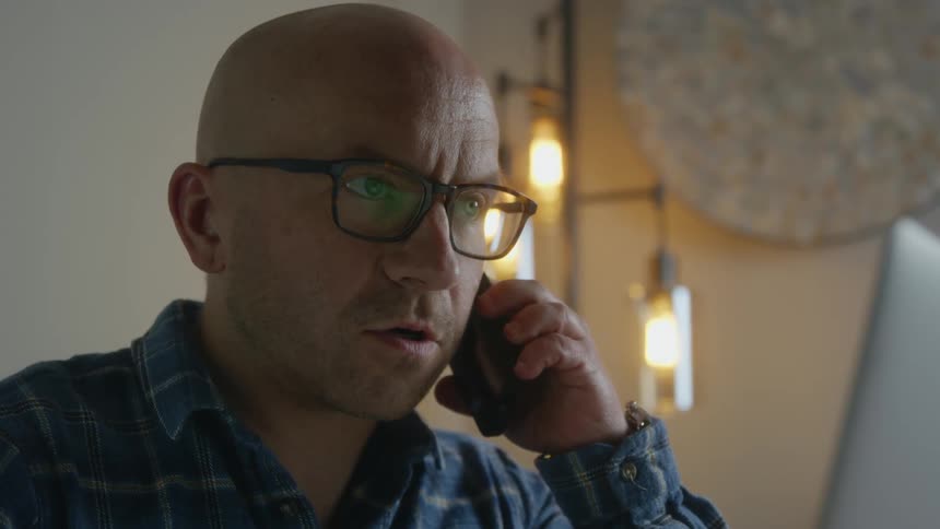 A man talks on the phone while sitting in front of his laptop