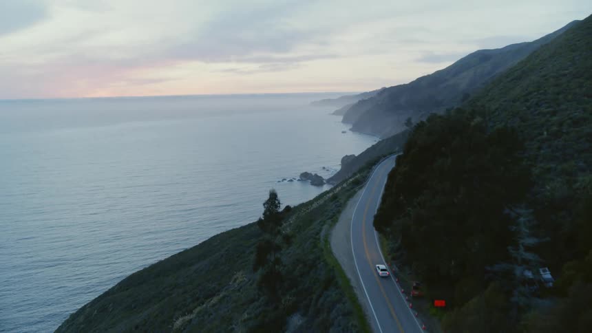 Car drives along winding mountainside road near Big Sur as red tints horizon