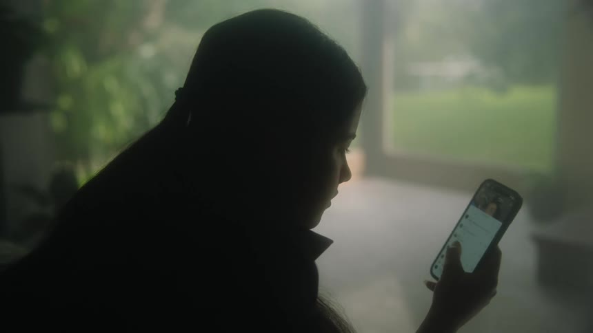 Woman using smartphone is silhouetted against the windows overlooking backyard