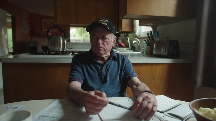Elderly man sits at table with paperwork looking around and tapping his pen