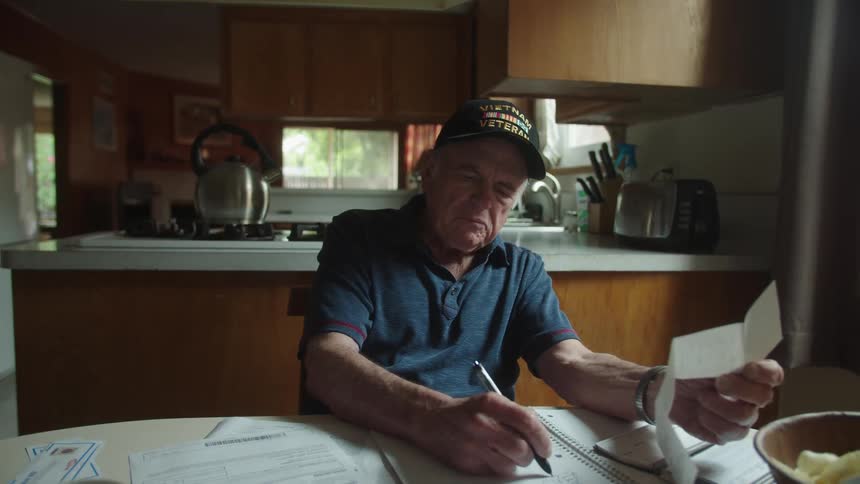 Elderly man sits writing budget information at table near kitchen