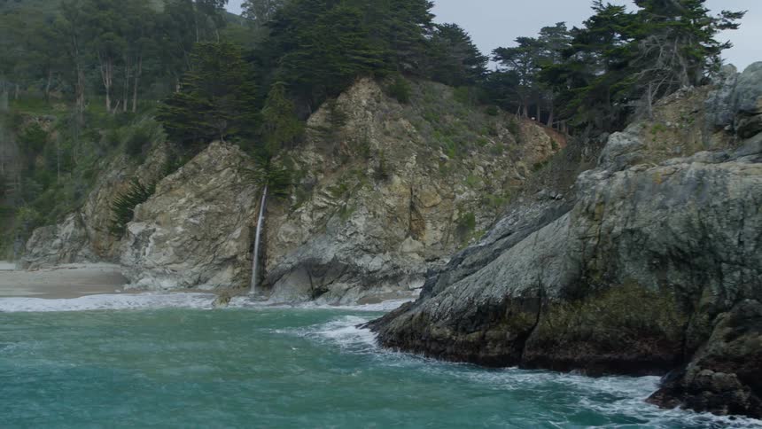 Narrow waterfall flows into ocean near beach at base of cliff topped with plateau