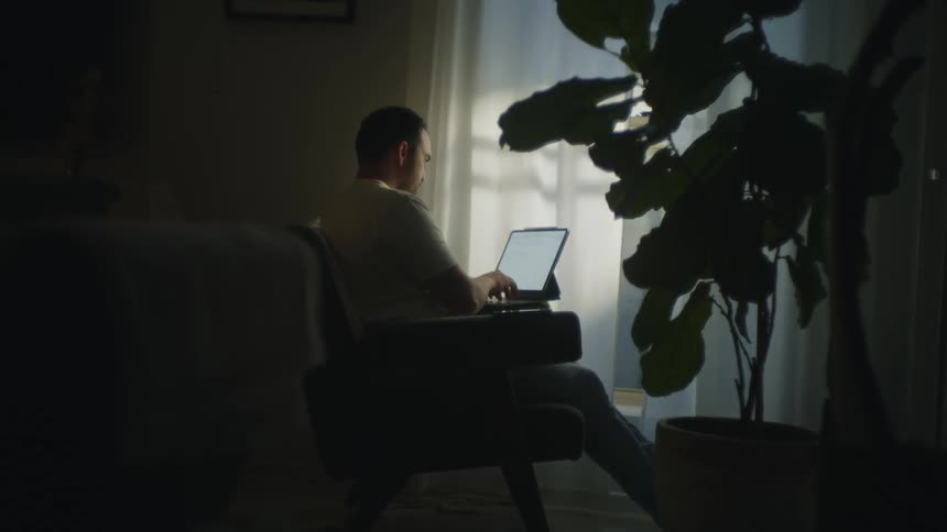 Man sits in dimly lit room typing on laptop in his lap
